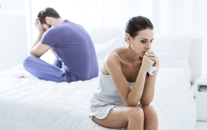 woman sitting on edge of bed with eyes closed, holding tissue. Man sitting on opposite side of the bed with hands on his face, looking down. Couple is contemplating marriage reconciliation after an infidelity.