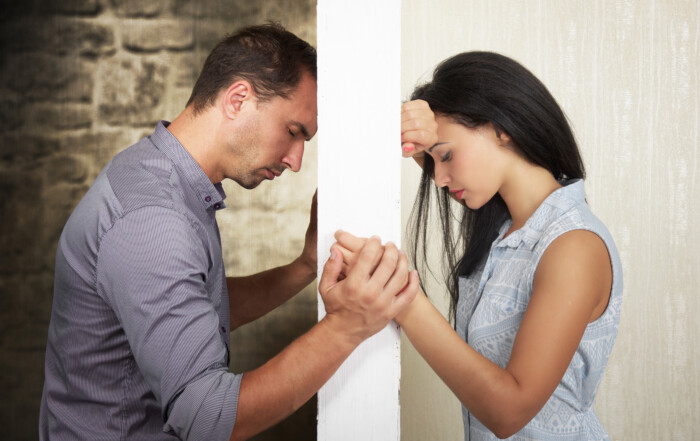 A man and woman with a wall separating them. Their hands come around the wall and are holding hands. They are both looking down, feeling sad. This couple is contemplating affair recovery counseling to save their marriage.